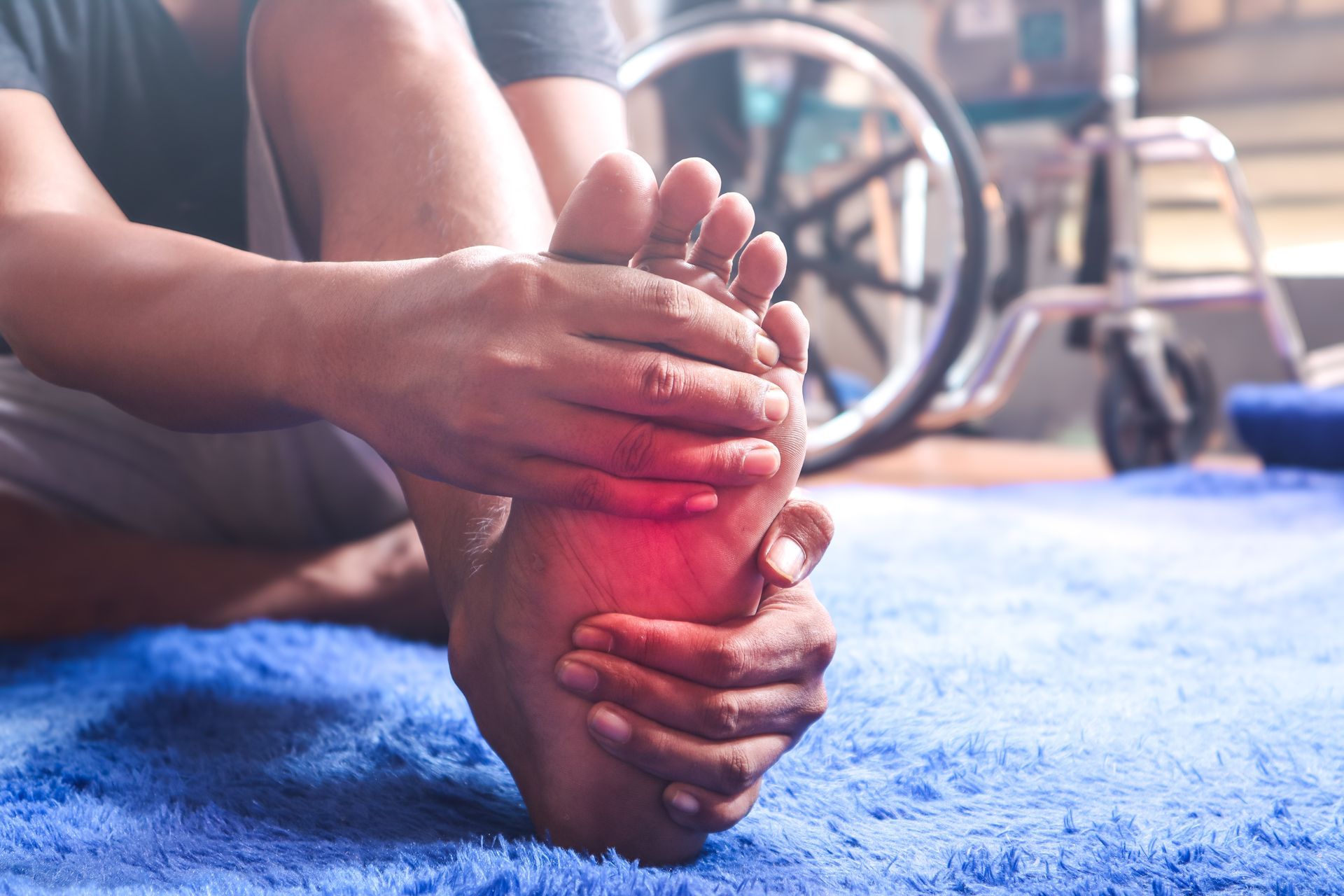 Person with red, swollen foot; holding foot, sitting on blue rug; wheelchair in background.