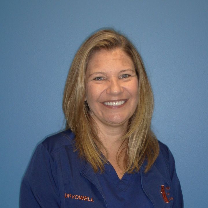 Woman with blonde hair in blue scrubs smiles at the camera, blue background.