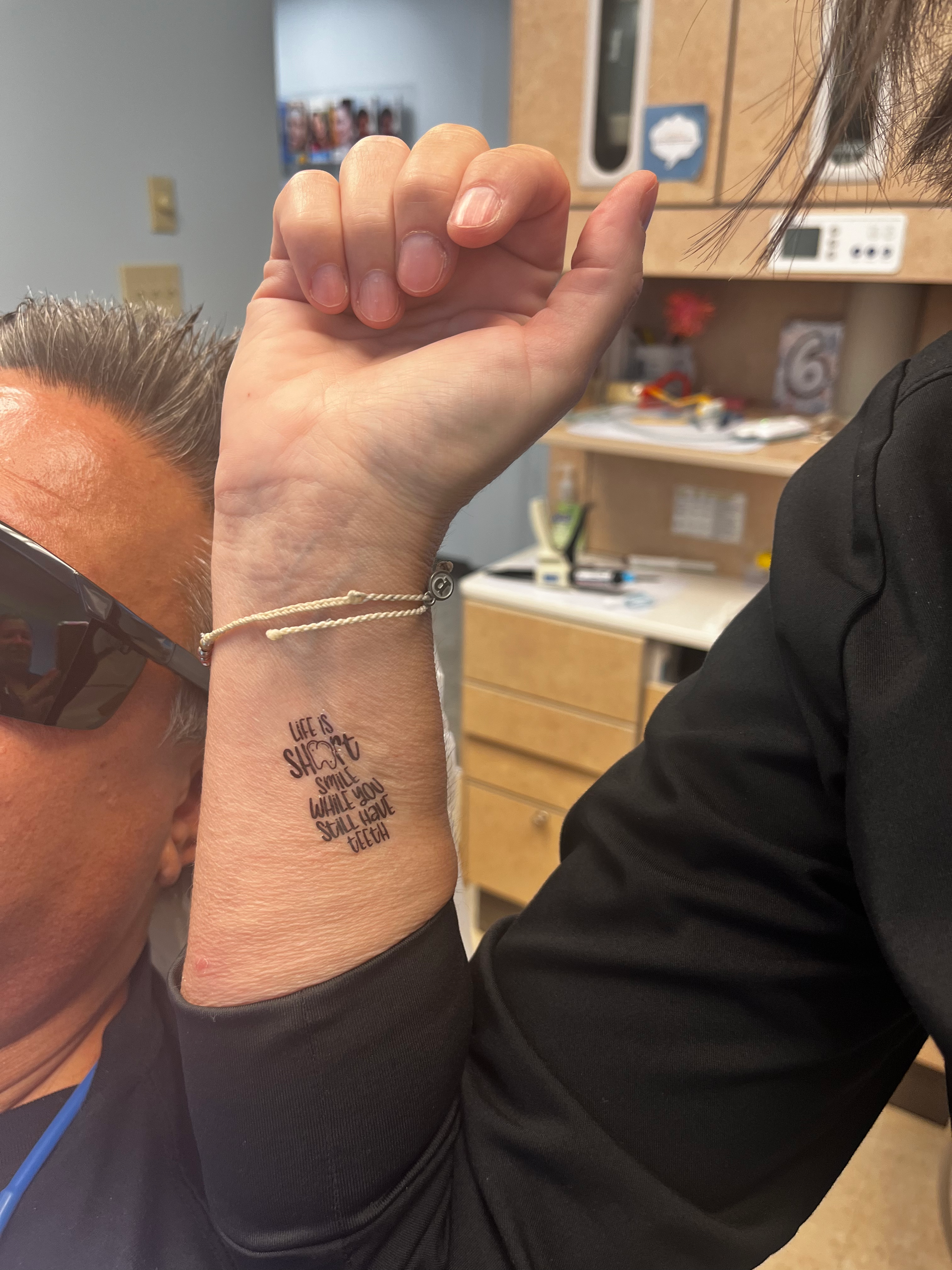 Person displaying a wrist tattoo; the tattoo is black and appears to be text. The person is in a medical office.