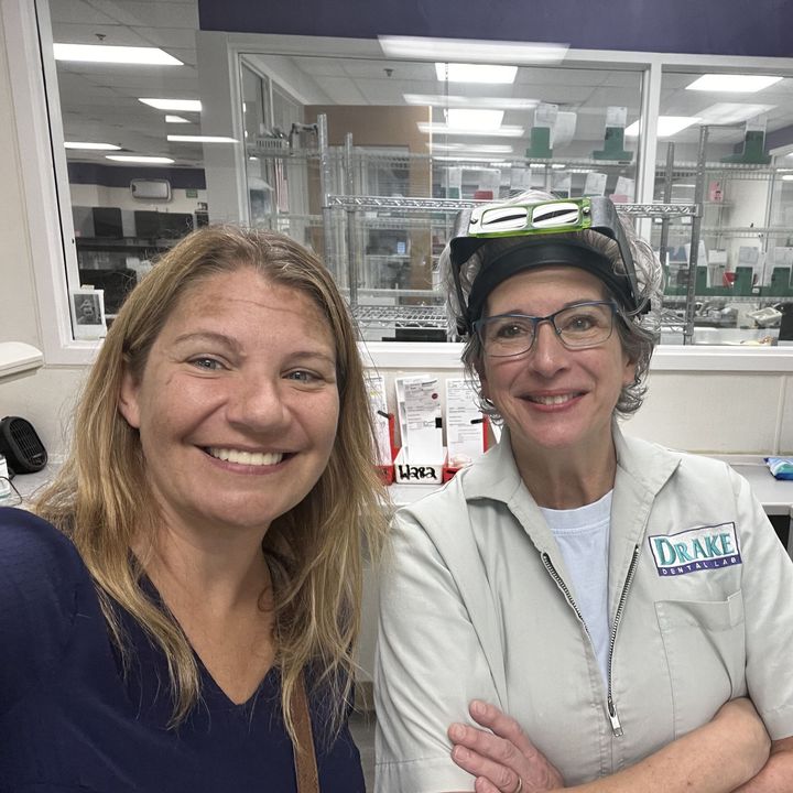 Two smiling women at a pharmacy counter; one wearing a Drake uniform and protective headgear.