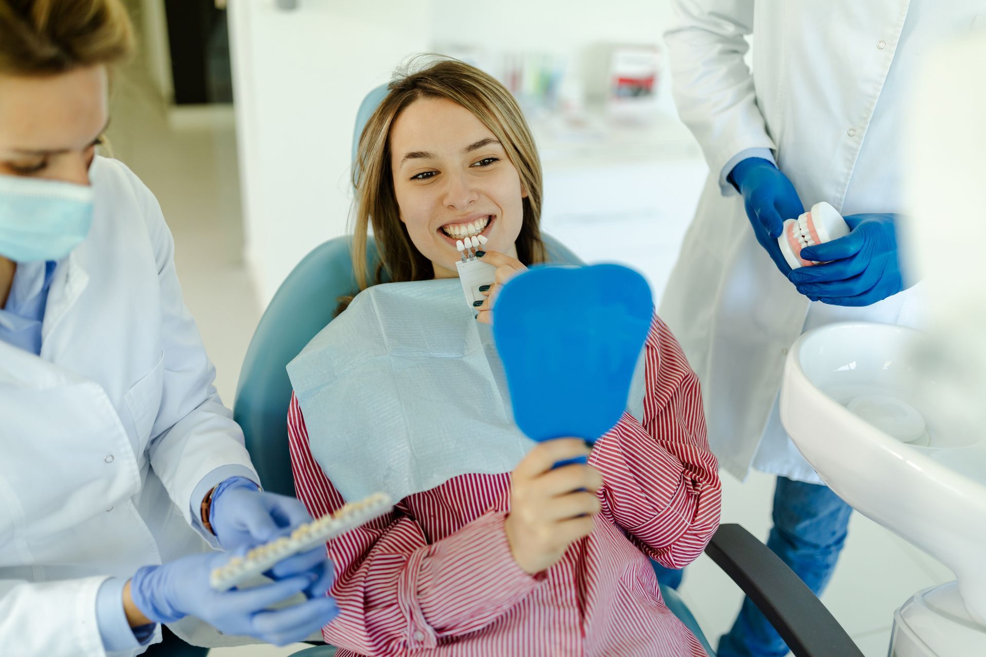 A woman is sitting in a dental chair looking at her teeth in a mirror.