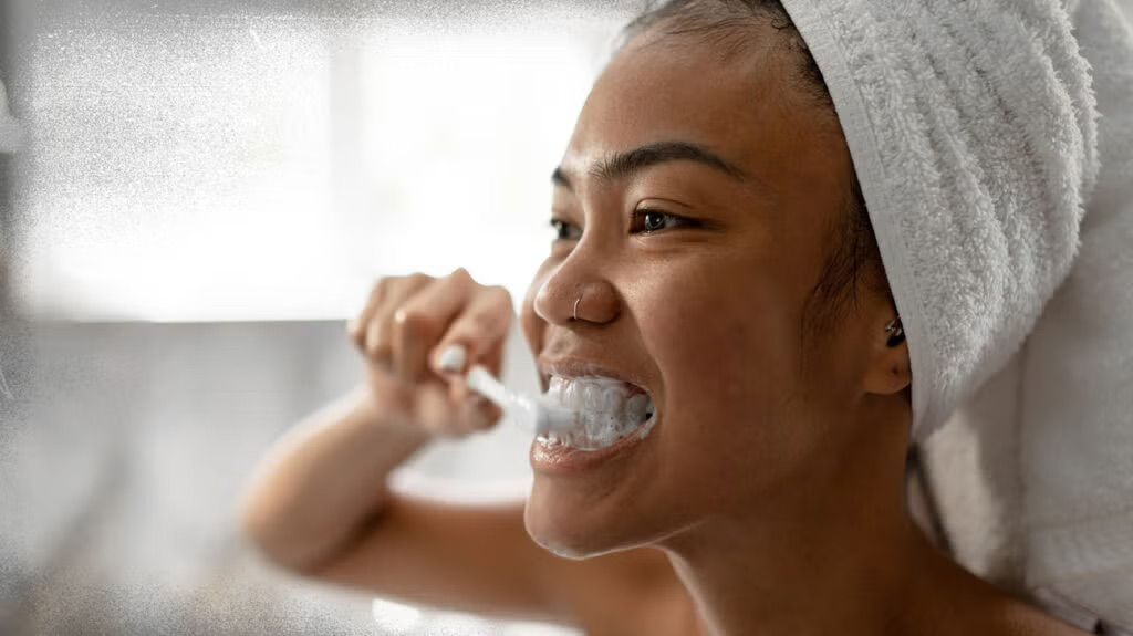Woman with towel on head brushing teeth, smiling.