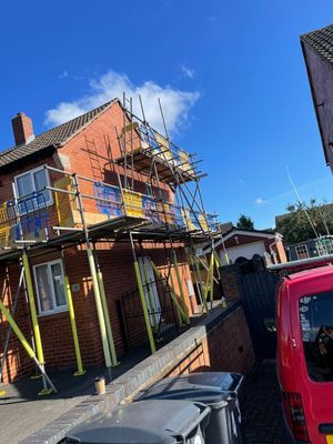 Scaffolding on brick house exterior, blue sky, trash cans, and red vehicle.