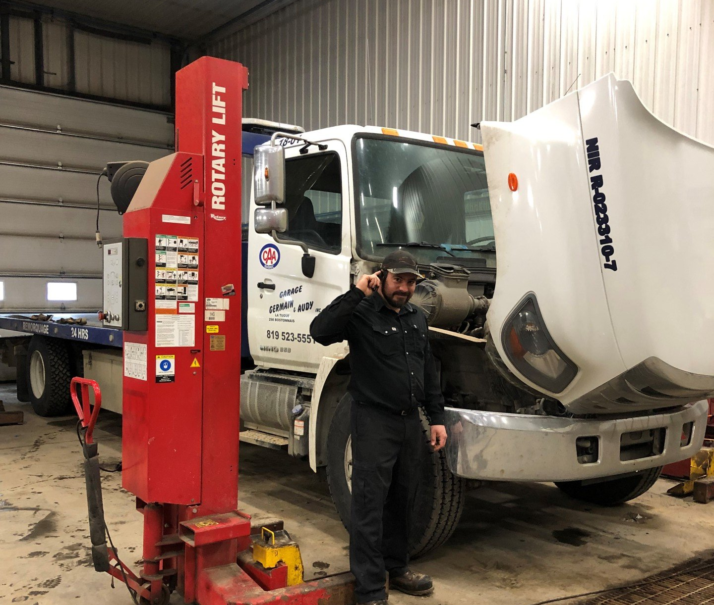 Dans un atelier, un mécanicien se tient à côté d'un camion dont le capot est ouvert, un pont élévateur rouge à côté.