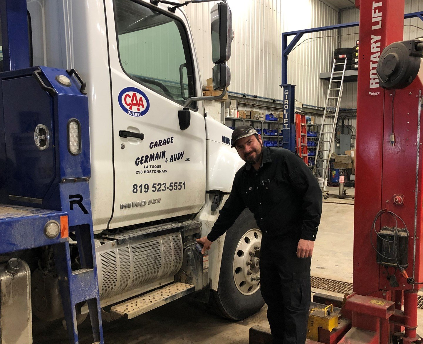 Un homme désigne du doigt le réservoir d'essence d'un camion, stationné dans un garage. Le camion est blanc et bleu, et l'homme sourit.