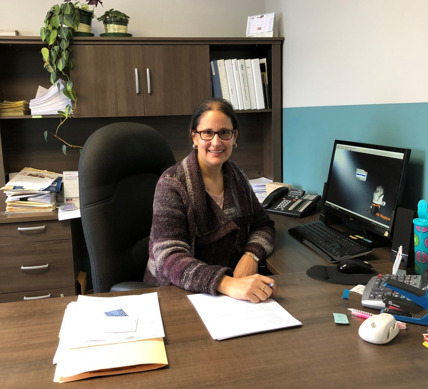 Une femme portant des lunettes est assise à son bureau, souriante. On aperçoit son ordinateur, son téléphone et des papiers.