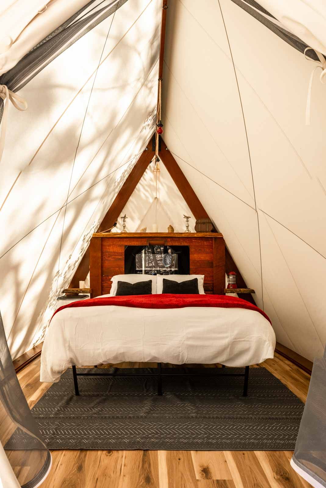 Interior of a canvas yurt tent with a bed, red bedding, and wooden headboard. A rug lies on a wooden floor.