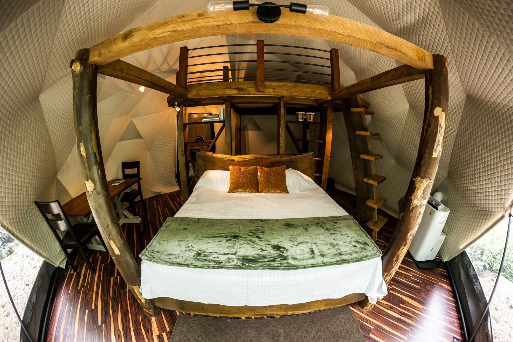 Inside a geodesic dome, a bed with a green coverlet and a loft above it, accessed by a ladder.