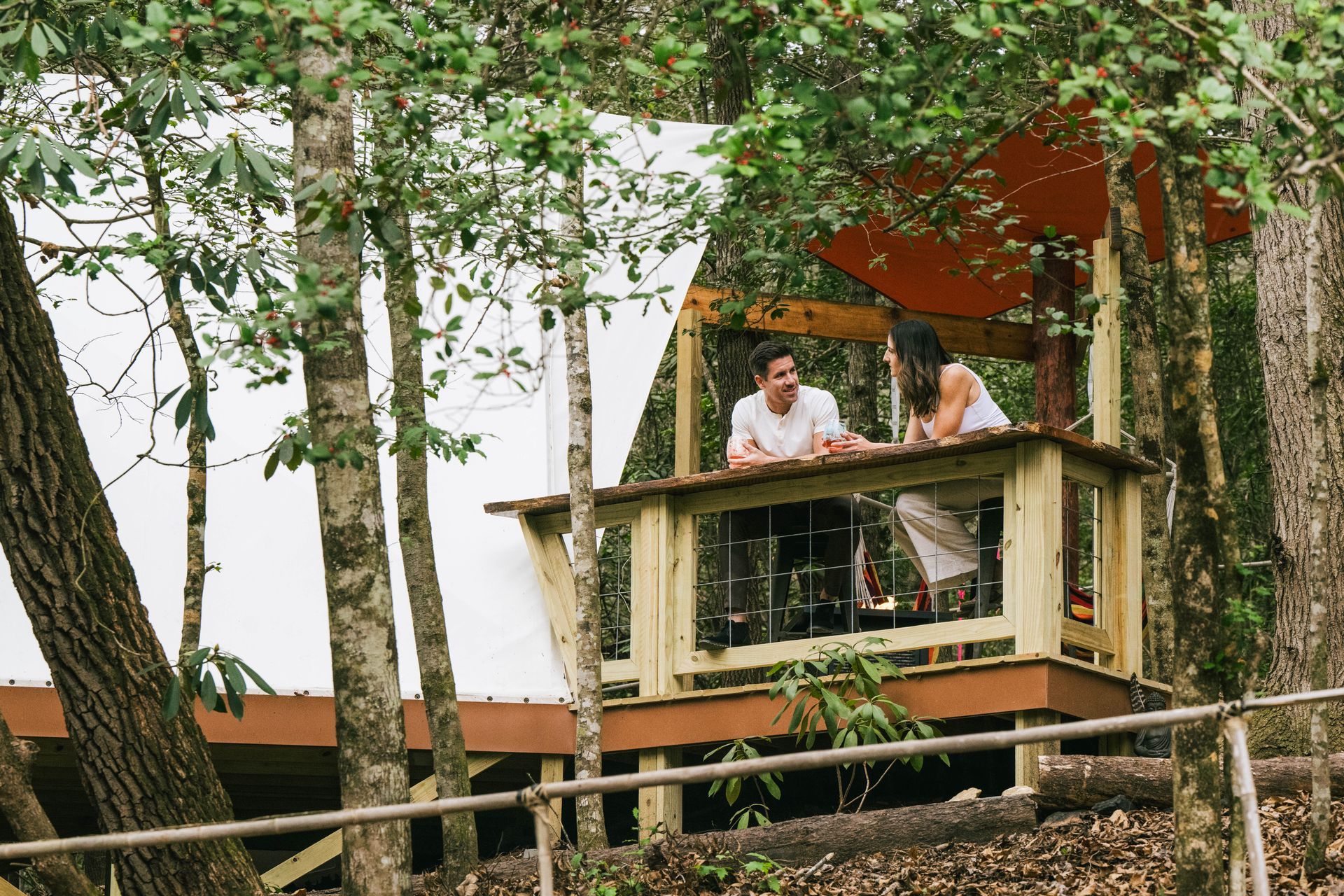 Two people on a wooden balcony under trees. White yurt tent structure visible, forest setting.
