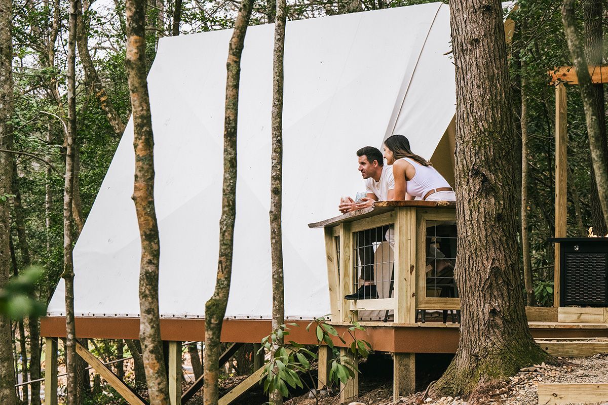 Couple on a cabin deck overlooking a forest. White tent structure on wooden platform.