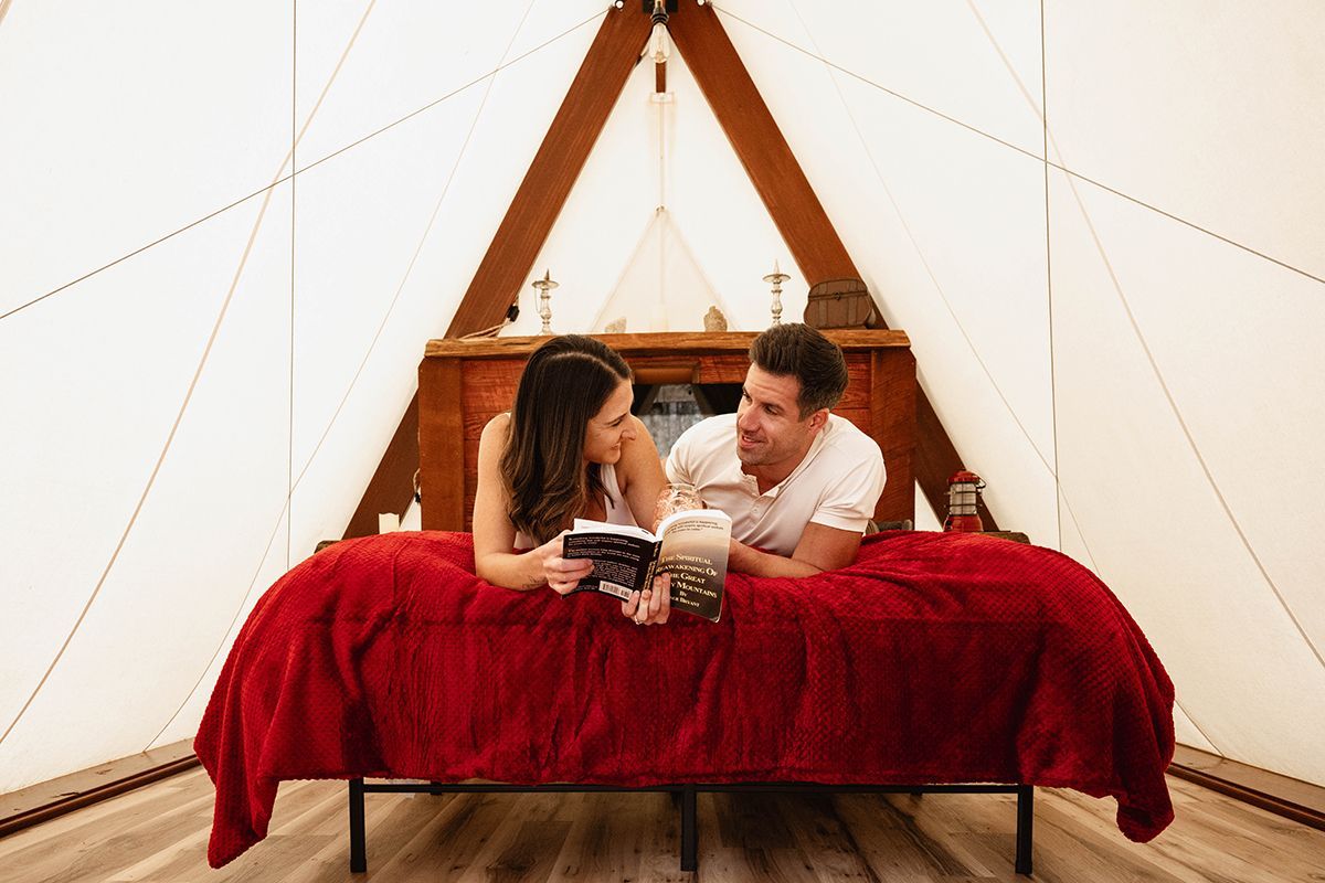 A couple lays on a bed reading a book in a tent.