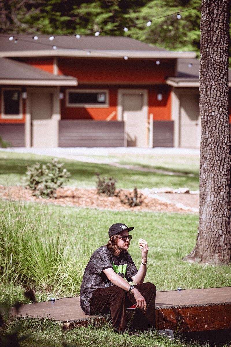 A man sits at a campground with a bath house in the background.