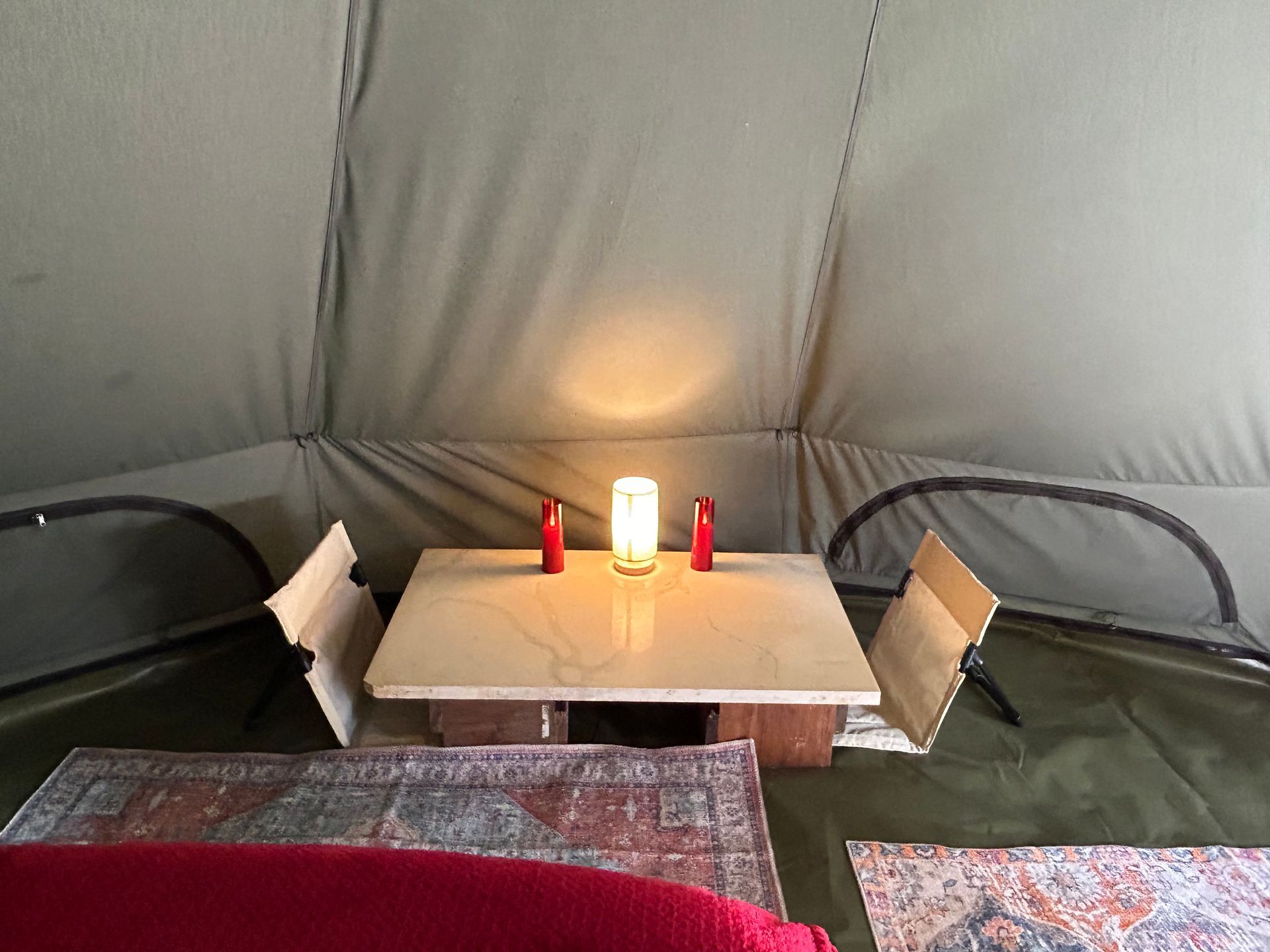 Inside a tent, a small table with a lit lamp and two red candles. Two cardboard panels flank the table, rugs below.