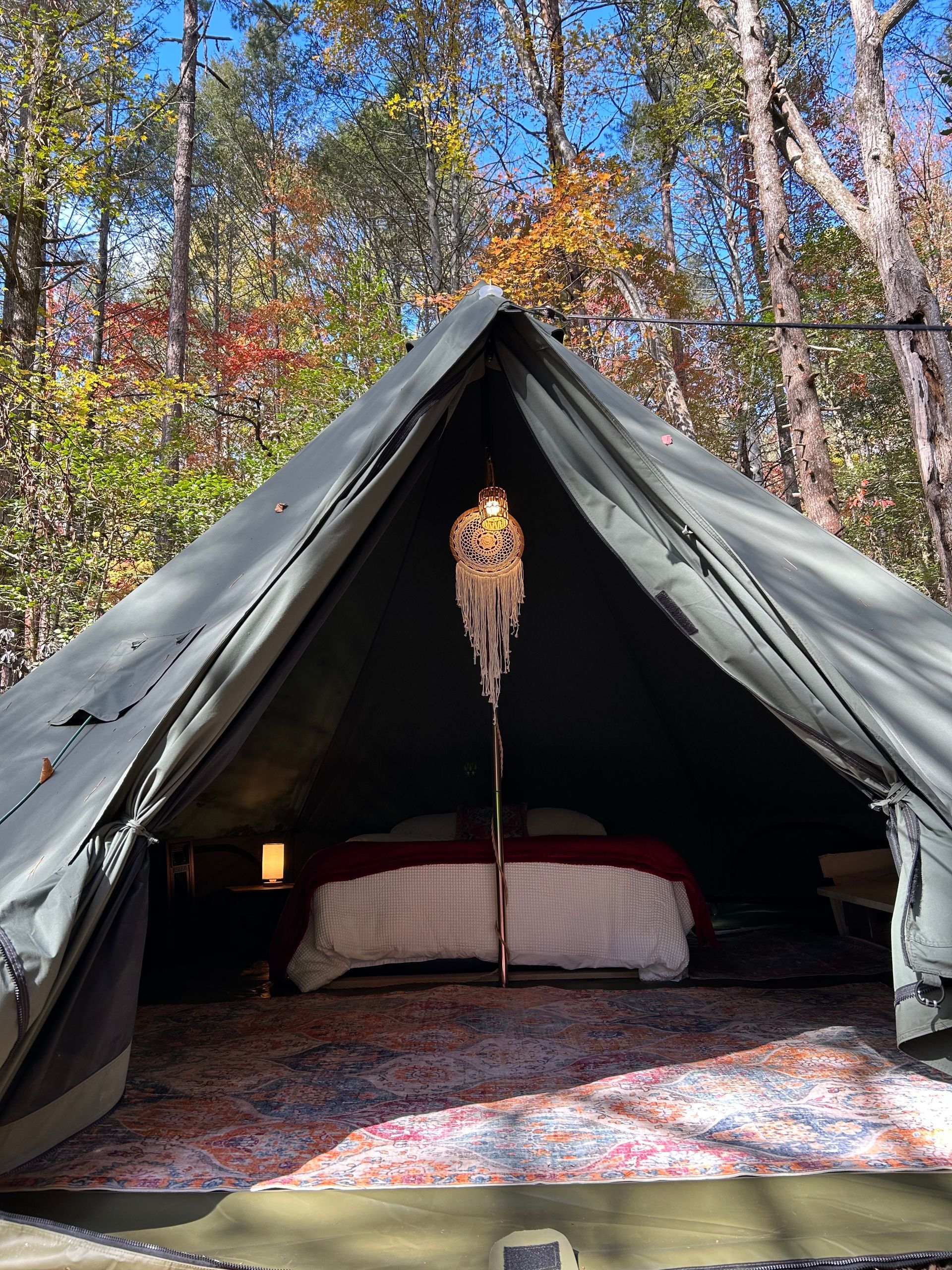 Large canvas tent interior with bed, rug, and hanging light, set in a forest.