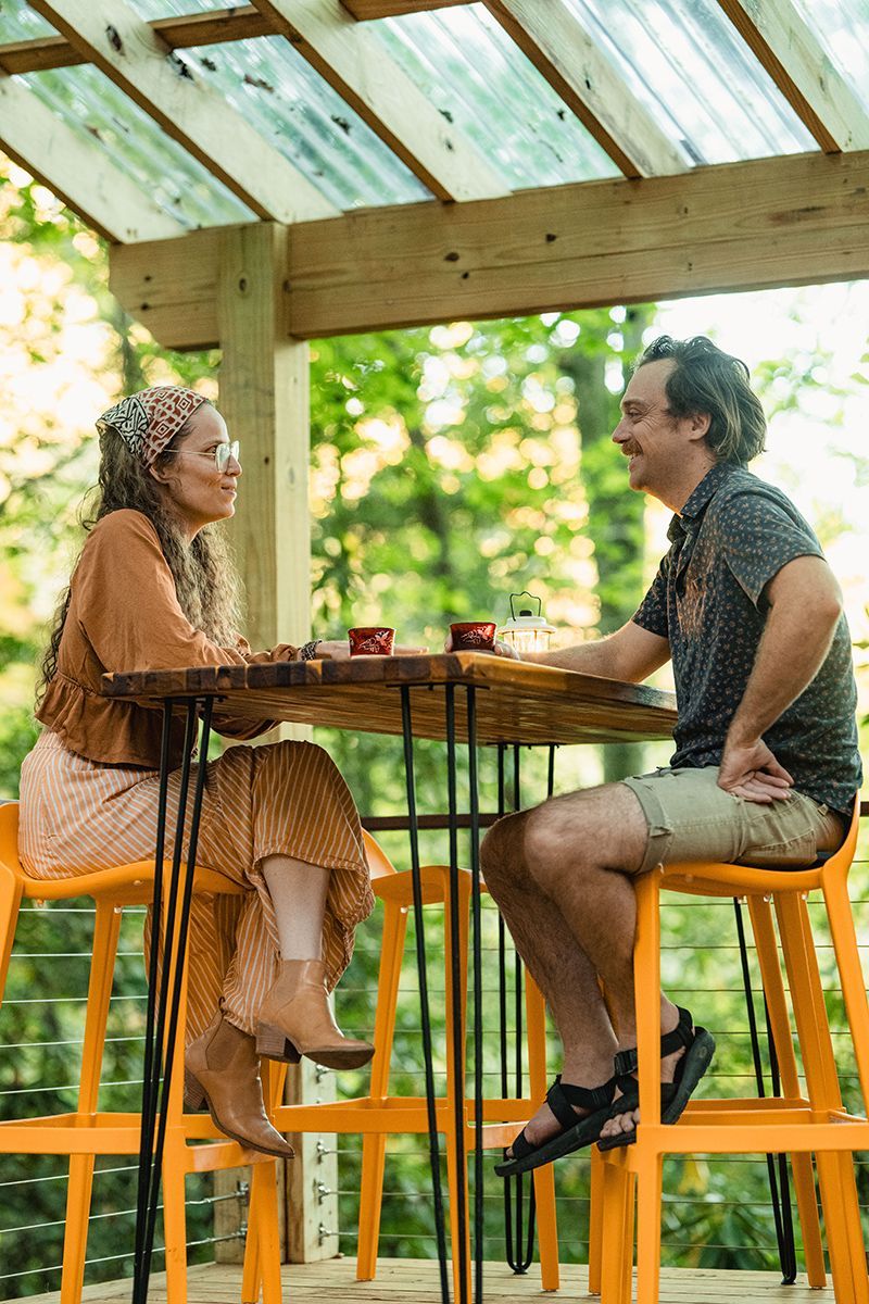 Couple seated at a high table, outdoors on creator cabin porch deck. Woman in orange, man in patterned shirt, smiling.