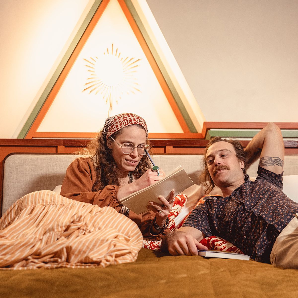 Couple in bed: one writing in a notebook, the other looking on, above a wooden headboard, under a triangle skylight.