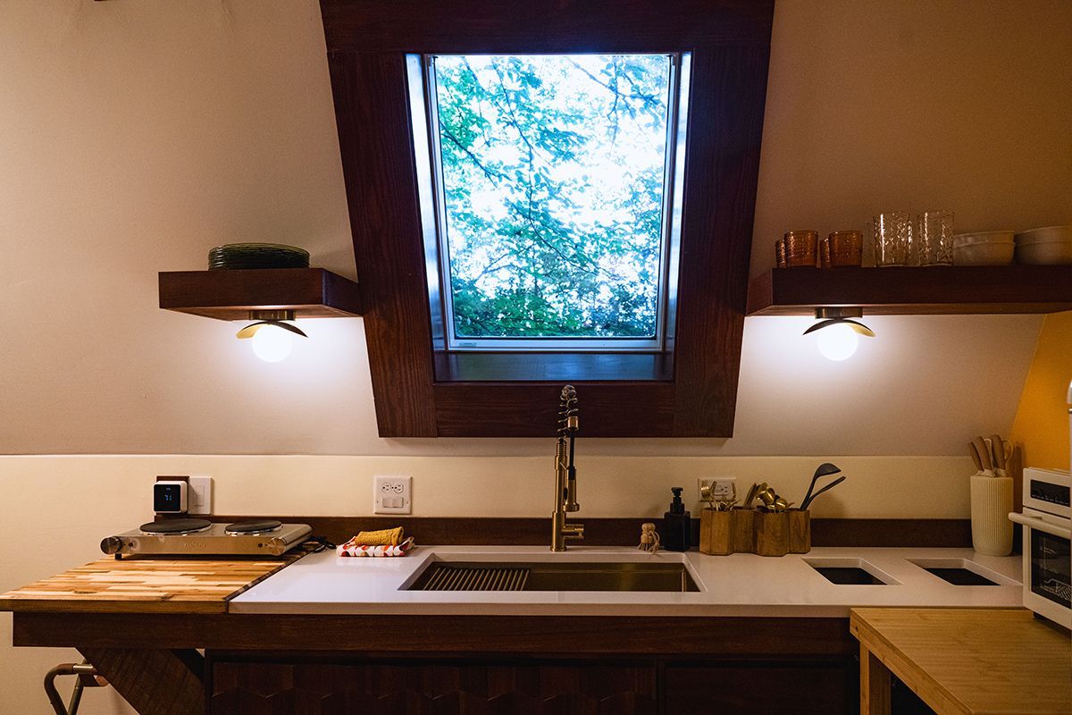 Small kitchen with sink, window above, shelves with lights. White countertop, wood accents, natural light.