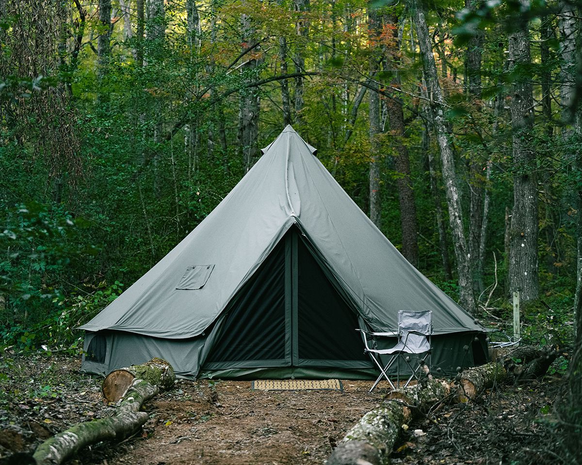 A canvas tent pitched in a forest clearing, with a folding chair outside.