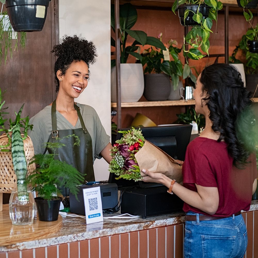 Flower shop clerk hands a bouquet to a smiling customer at the counter, with hanging plants and potted greenery.