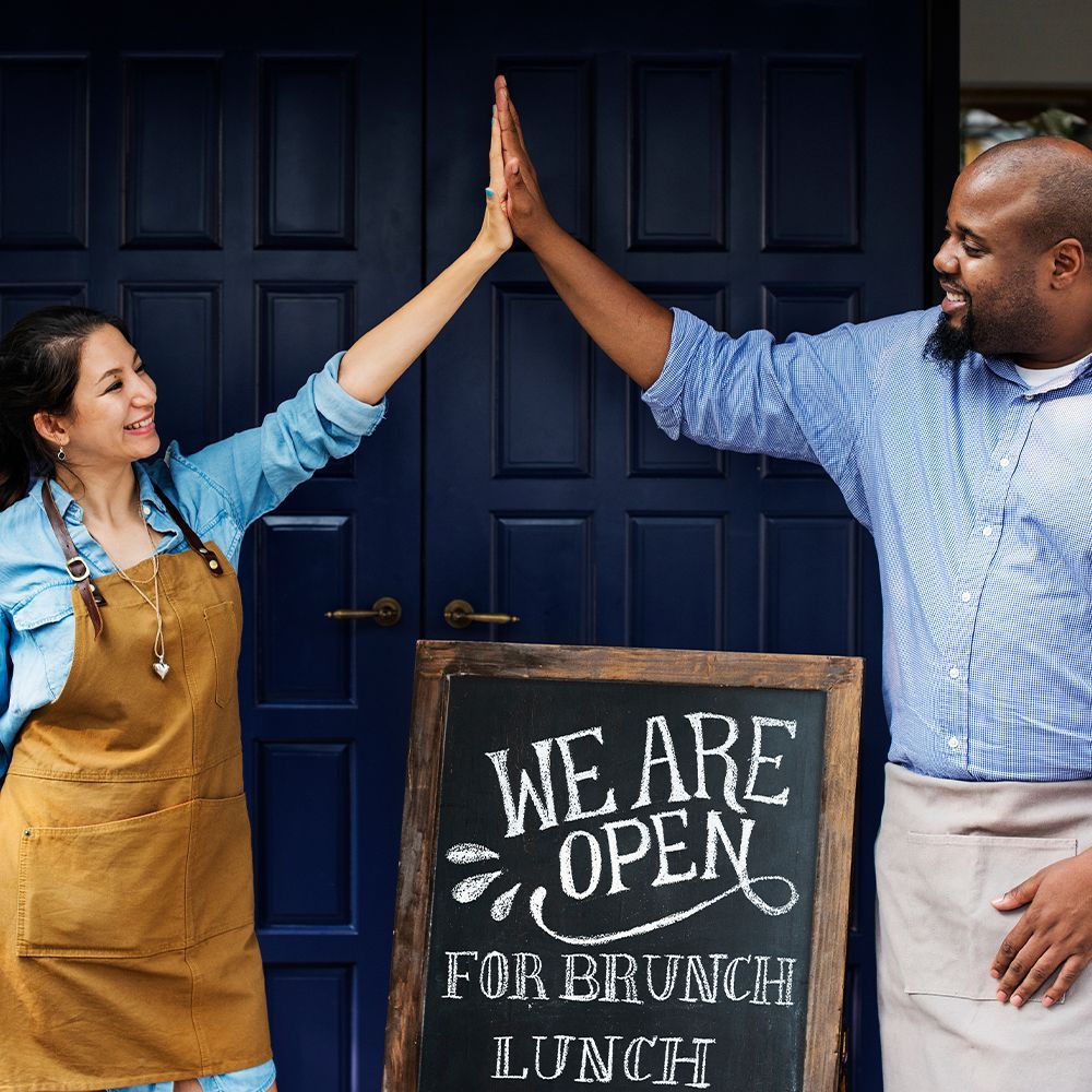 Two coworkers high-five in front of a blue door and “We are open for brunch” sign.