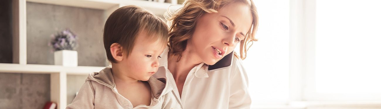A woman in a white shirt holds a toddler, with the child looking away. Bright, natural light.