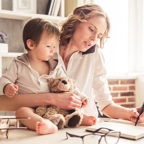 Woman works at a desk, holding a child with a teddy bear. Both are indoors, in a well-lit room.