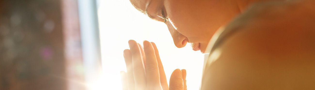 A person with their head down, hands touching a bright window, bathed in sunlight.