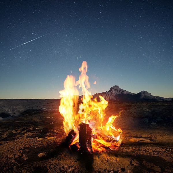 Campfire blazing under a starry night sky, with a mountain in the background.