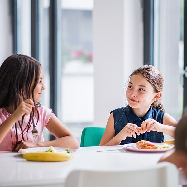 Two young girls eating at a table, smiling and talking in a bright cafeteria.