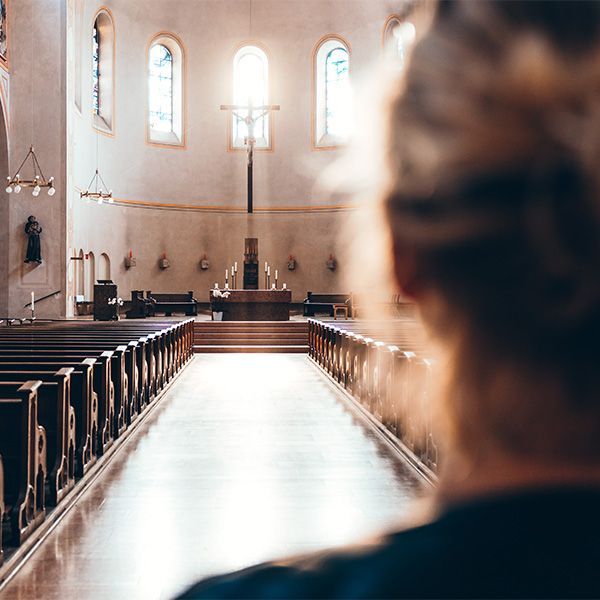 Woman facing the aisle of a sunlit church, wooden pews, altar with a crucifix.