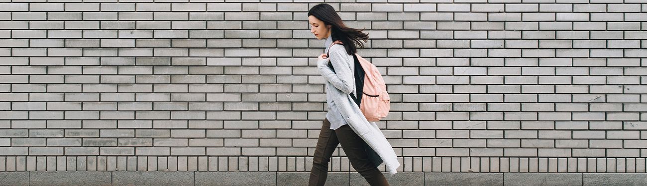 A woman with a backpack is walking in front of a brick wall.