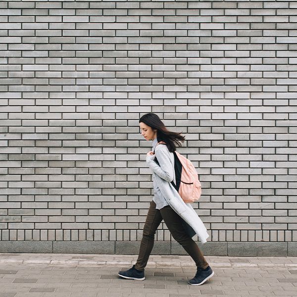 A woman with a backpack is walking in front of a brick wall.