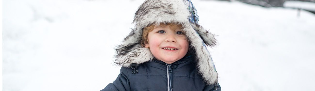 A young boy with a big fur hat smiles in the snow.