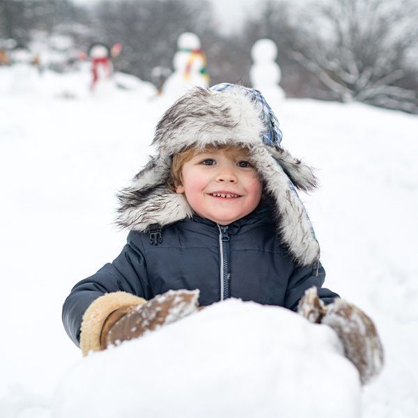 Young child smiles while rolling a snowball in snowy scene, wearing fur hat and mittens.