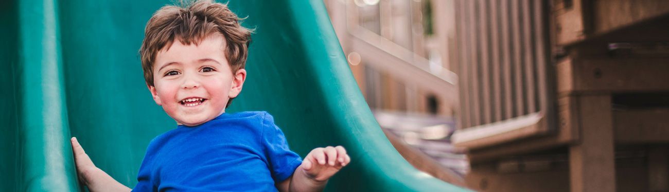 A young boy with curly brown hair smiles while sliding down a green slide in a playground.