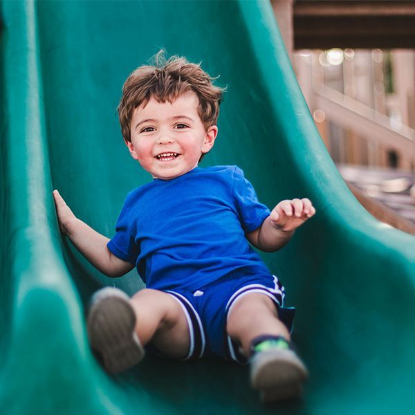 Happy young boy sliding down a green slide at a park, wearing blue and smiling.