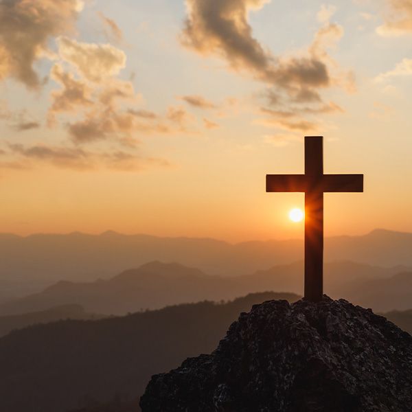 Cross silhouette atop a mountain peak at sunset with orange sky.