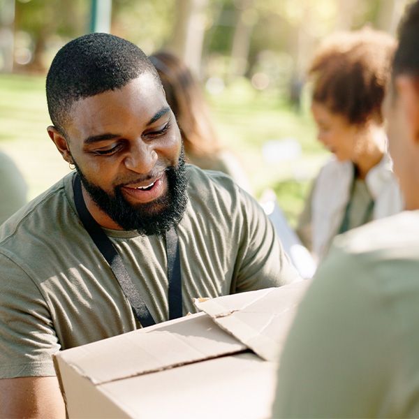 A man with a beard is holding a cardboard box and smiling. He is volunteering.