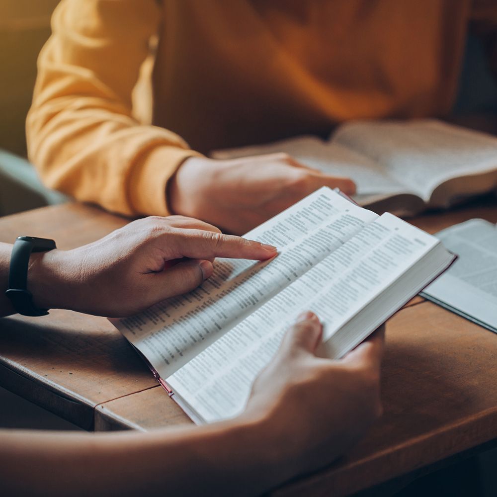 People reading open Bibles at a table, with a close-up of hands turning pages.