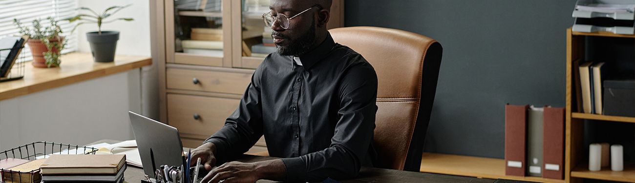 Parish priest typing on a laptop in an office.