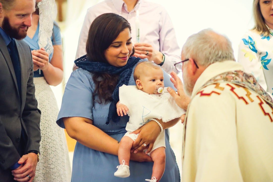 A woman holding a child while a Priest blesses her.