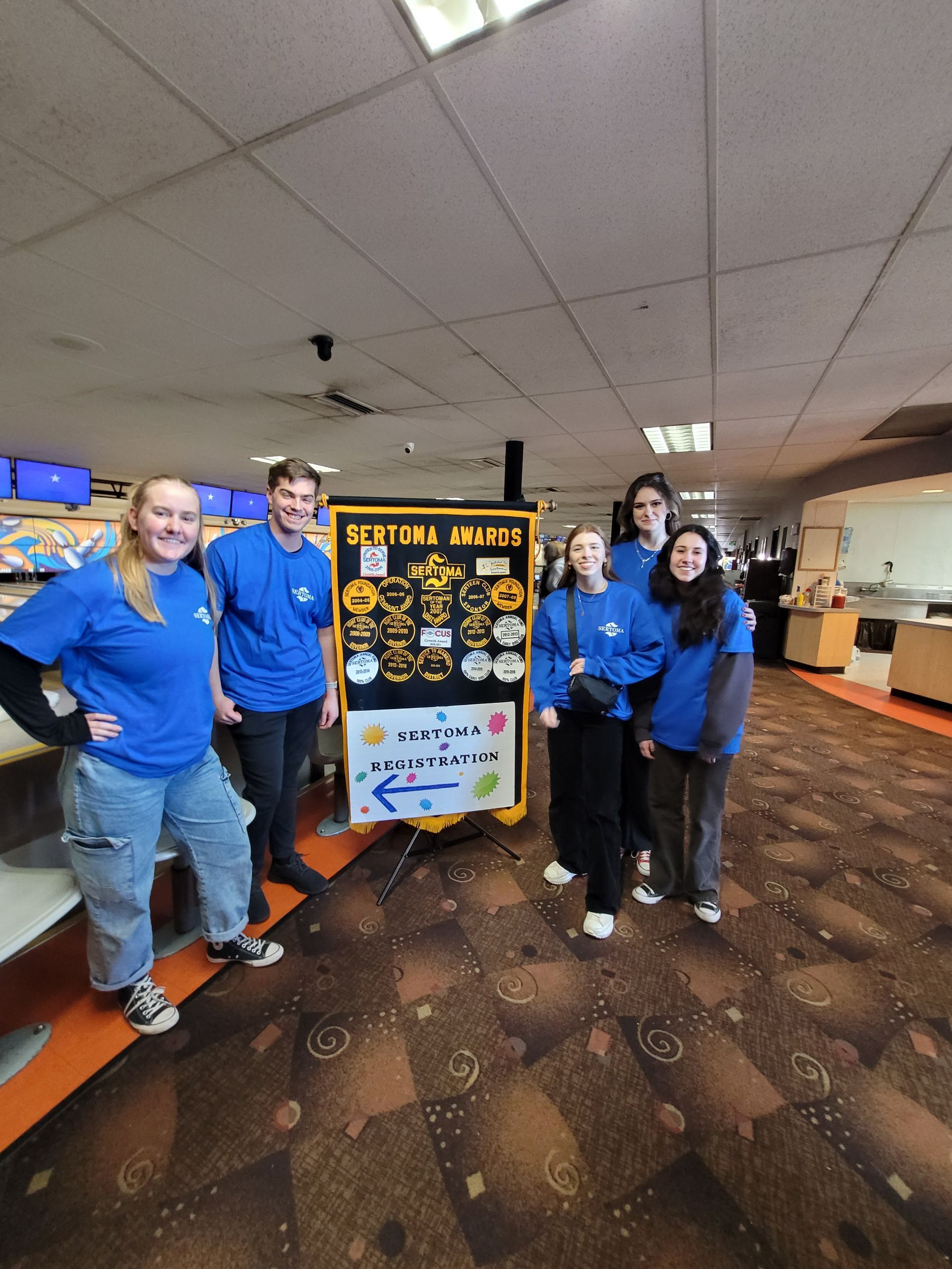 A man in a blue shirt is standing in a bowling alley talking to a group of people.