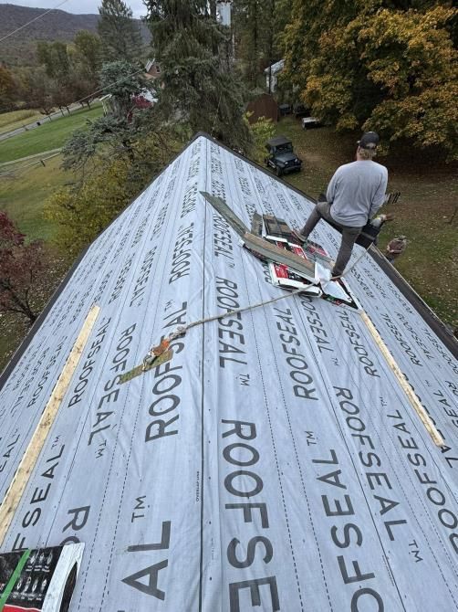 Roofer on a roof covered in "ROOFSEAL" underlayment, installing shingles. Green trees and a vehicle in background.