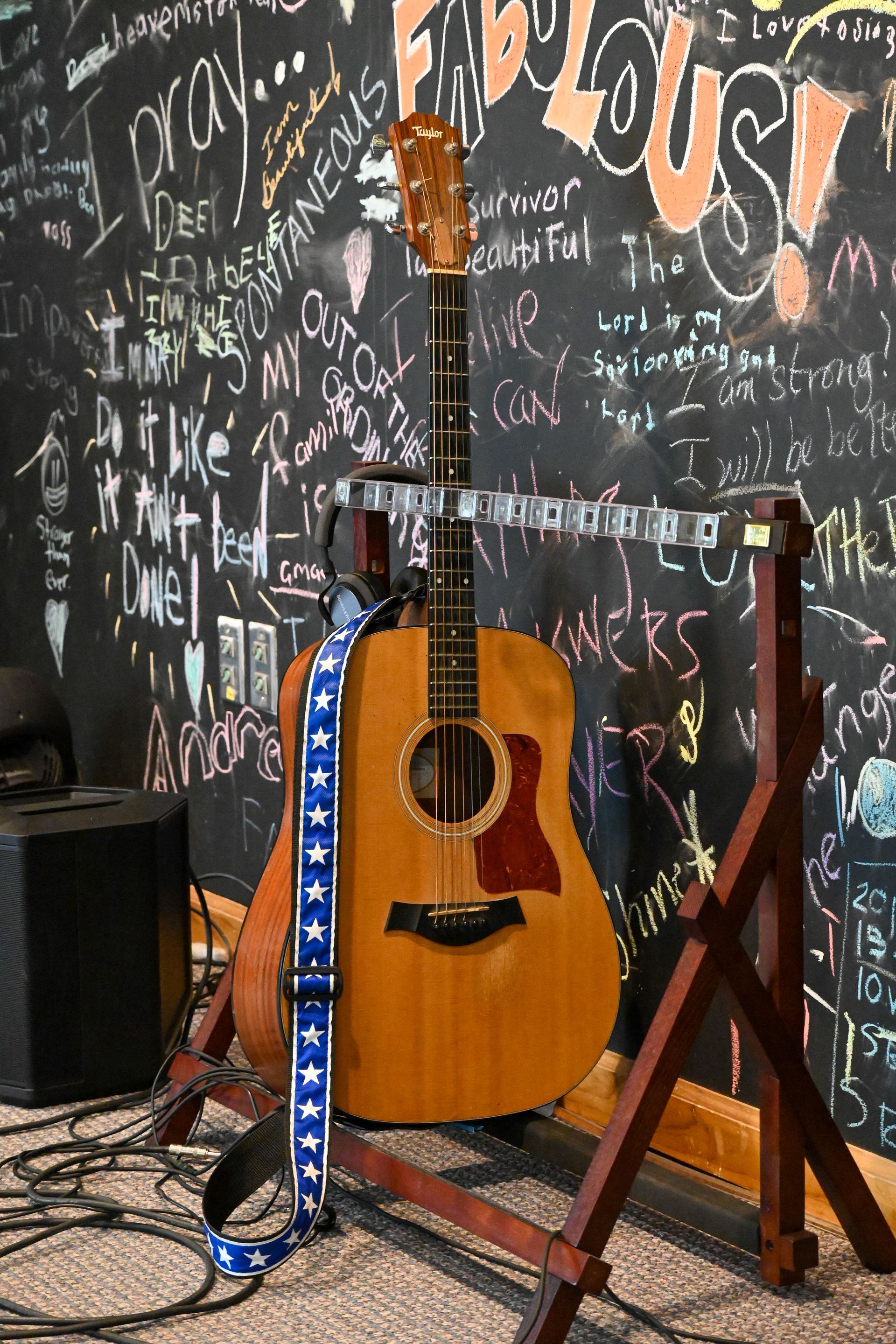 An acoustic guitar is sitting on a stand in front of a chalkboard wall.