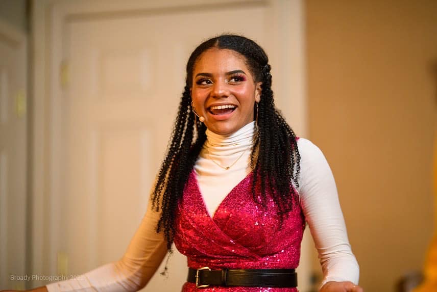 A woman in a pink dress and white turtleneck is smiling.