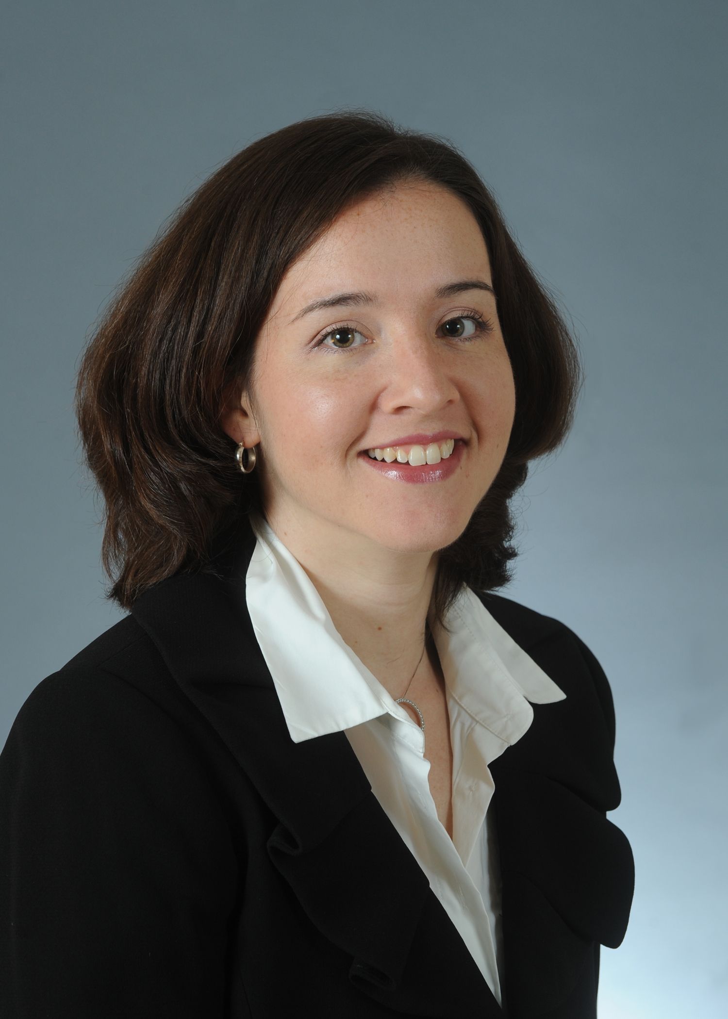 A person with shoulder-length dark hair wearing a black blazer and white shirt, smiling against a gray background.