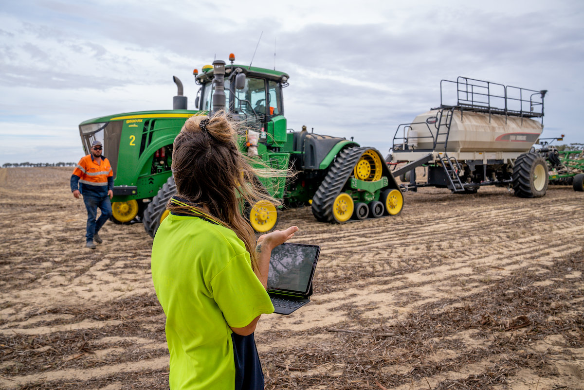 Woman using tablet to monitor a tractor and attached trailer in a field. Green, yellow, and brown colors are prevalent.
