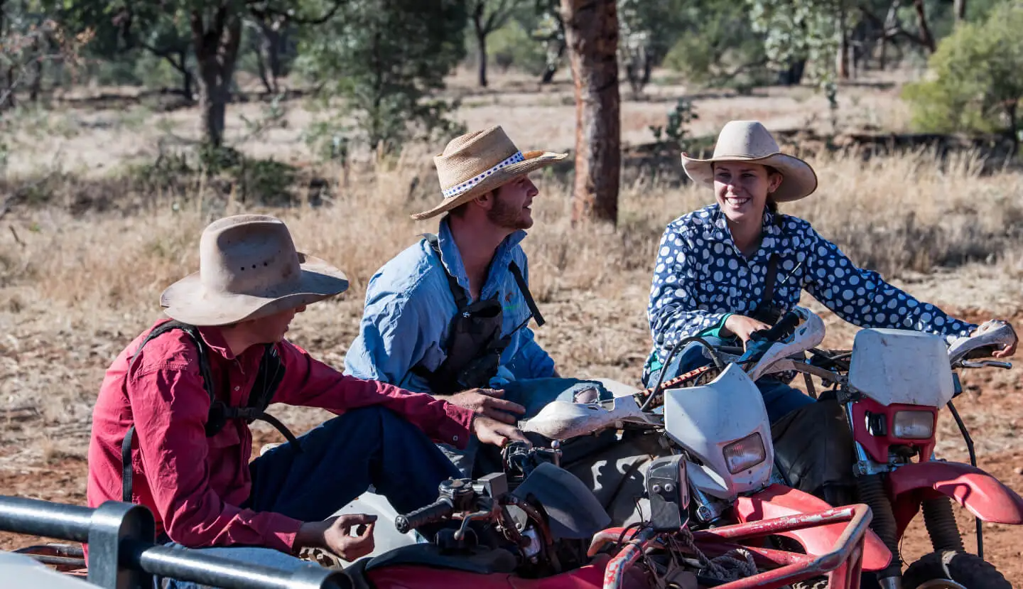 Three people wearing hats on a red ATV in a sunny, wooded area; they are smiling and chatting.