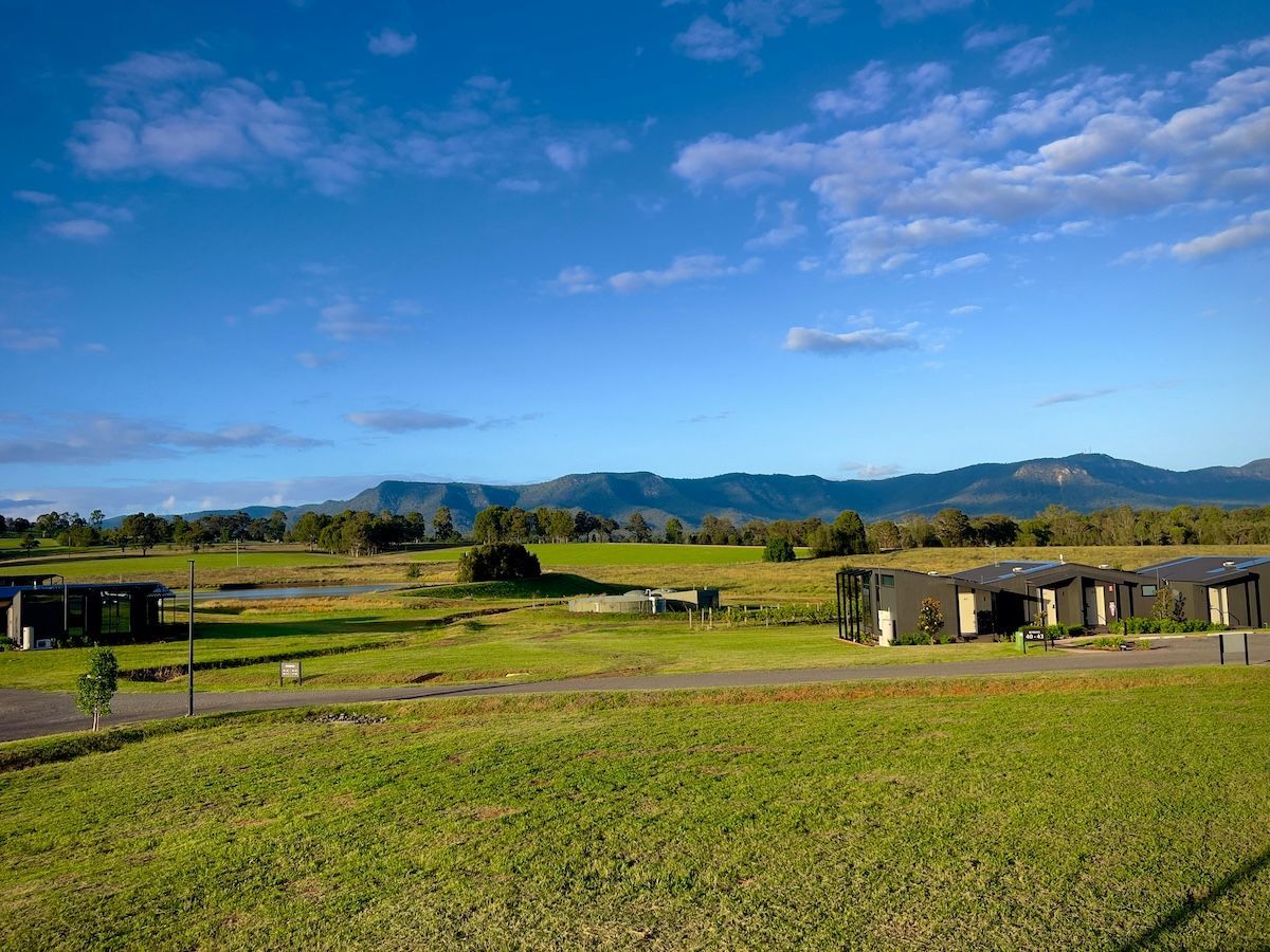 Grassy field with low-lying modern homes under a blue sky with mountains in the background.