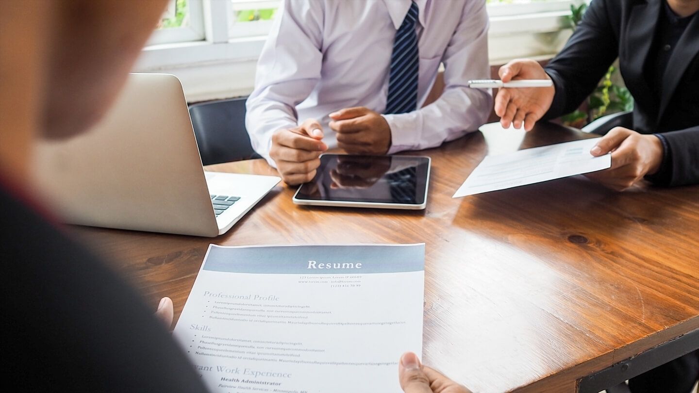 Person holding a resume at a table, meeting with two people, one holding a pen, and a laptop and tablet.